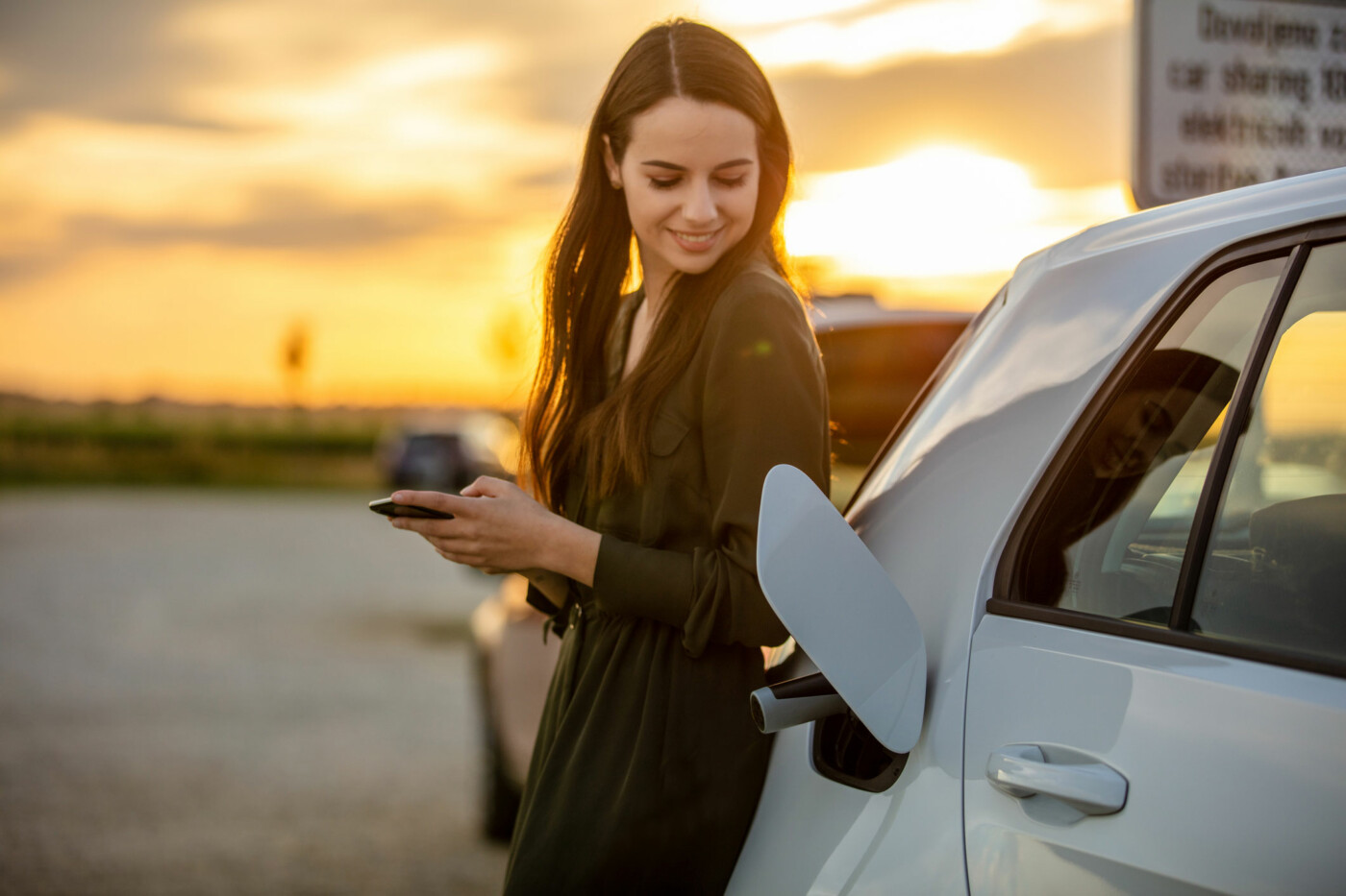 Eine Frau in einem grünen Kleid steht neben einem Auto und schaut auf ihr Smartphone während des Sonnenuntergangs.